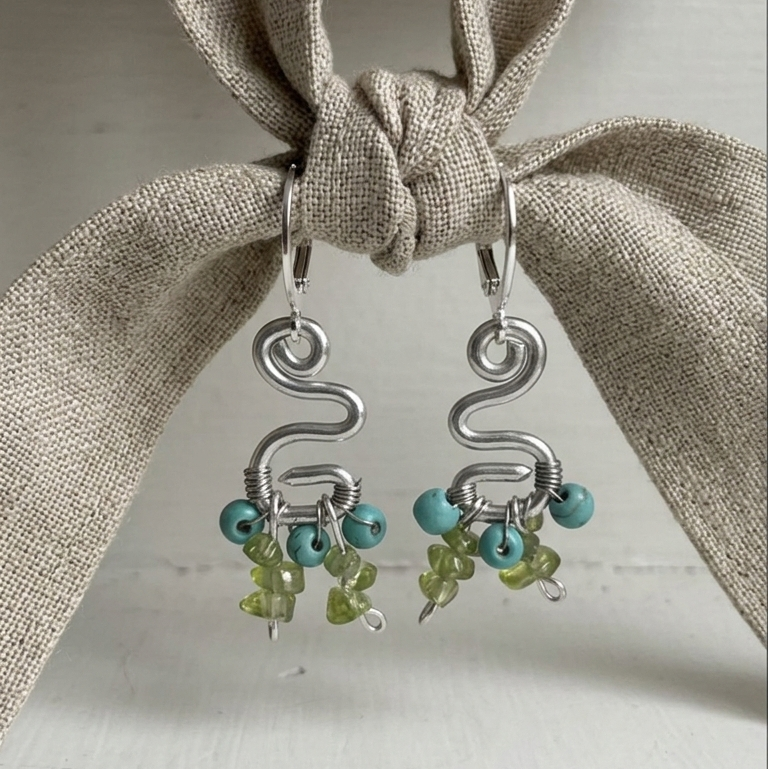 Silver earrings with turquoise and green beads hanging on a beige fabric background.