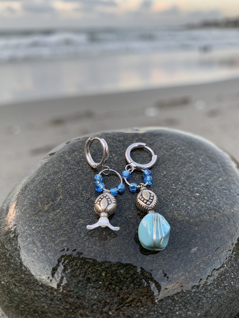 Two silver earrings with blue beads and charms on a rock by the water.